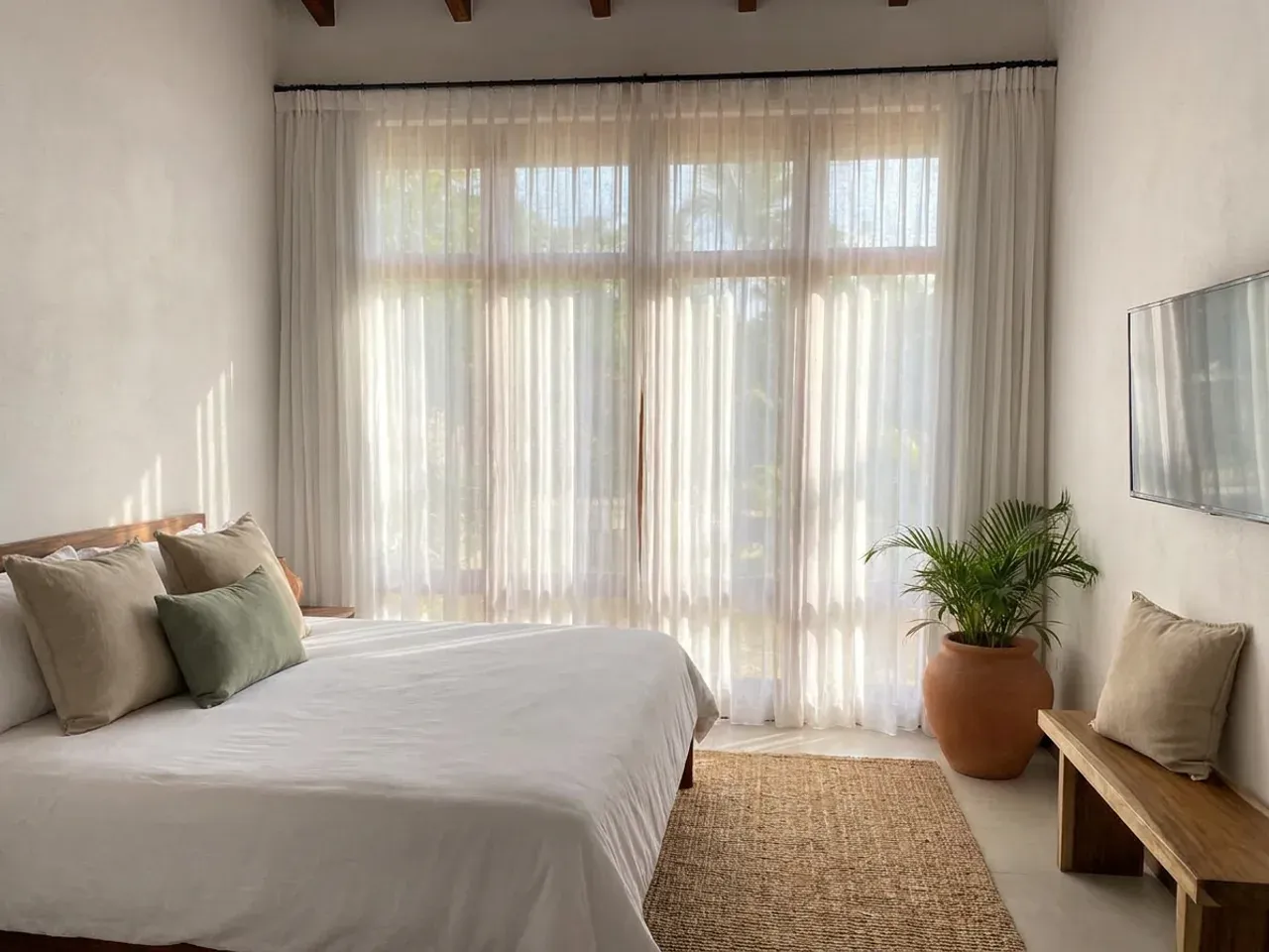 Dormitorio con cortina de tela clara y velo corridos sobre el ventanal, cama con ropa de cama de lino blanco, alfombra de fibra natural y planta en maceta de barro.