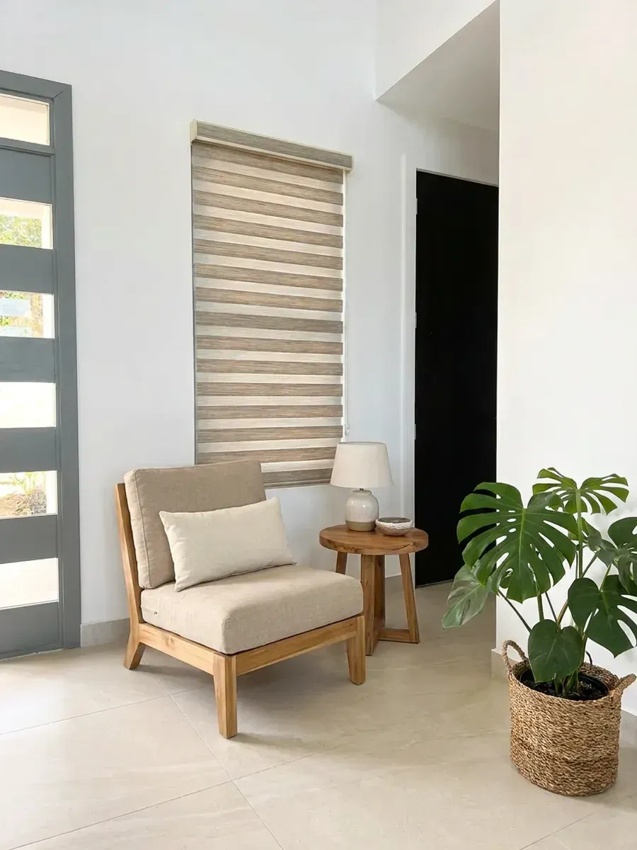 Double Roller bajado junto a una puerta de vidrio, en un rincón de estar con butaca de madera y planta.