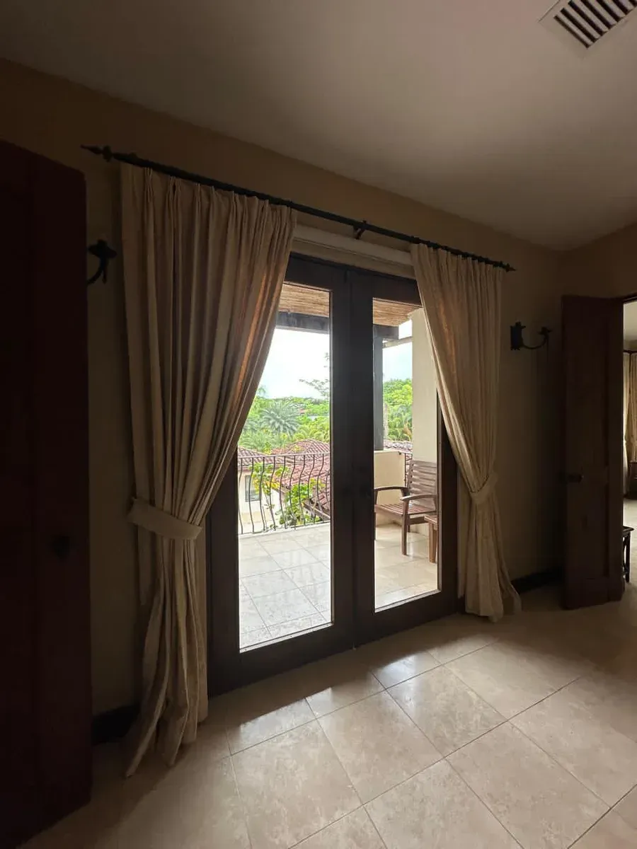 Off-white pleated drapes framing dark French doors with a balcony and tropical greenery beyond.