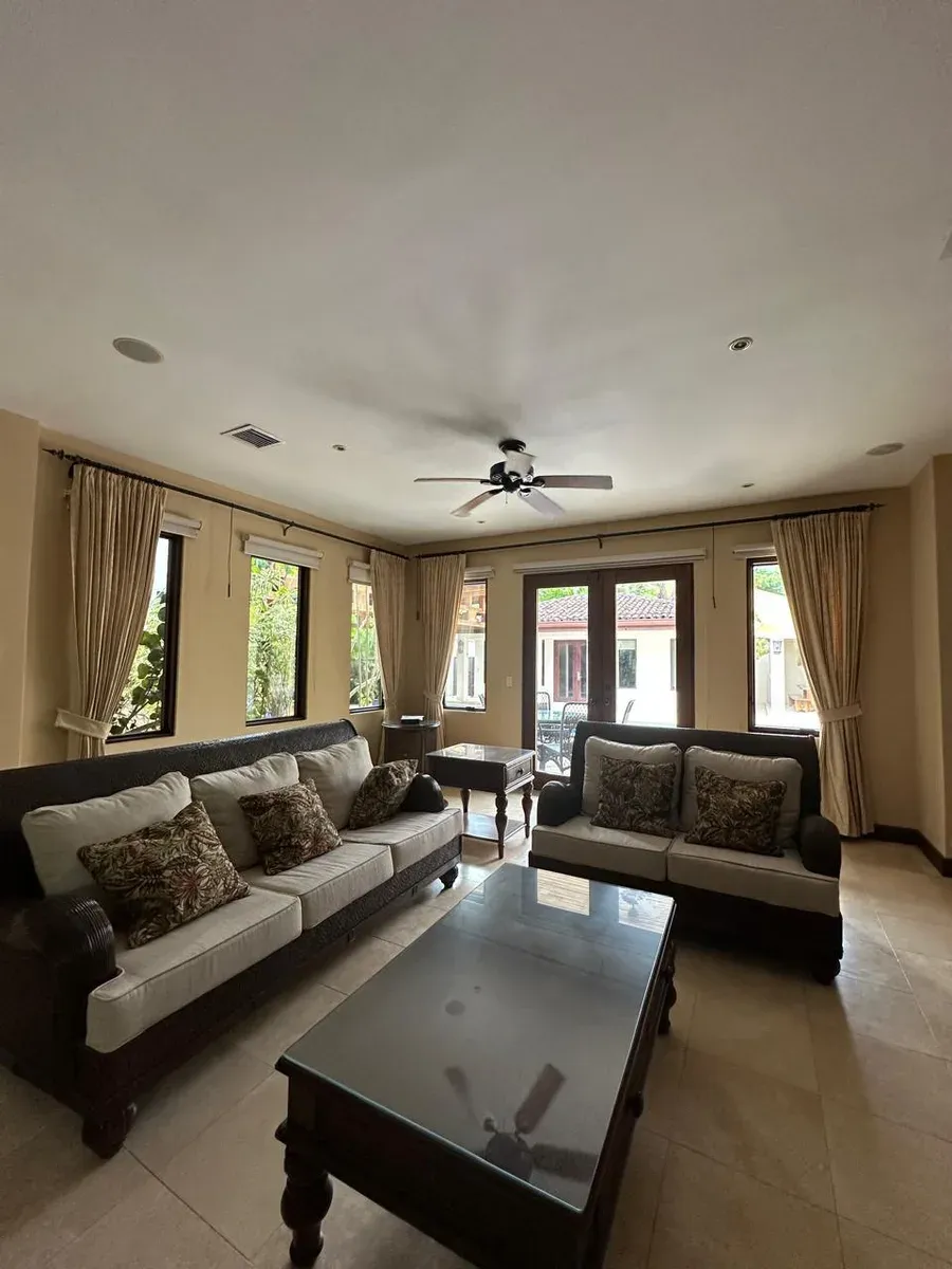 Living room with patterned sofas, ceiling fans, and cream pleated drapes across windows and doors.