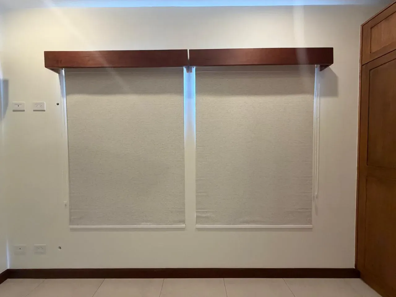 Two cream screen roller shades beneath dark-wood pelmets on a white wall.
