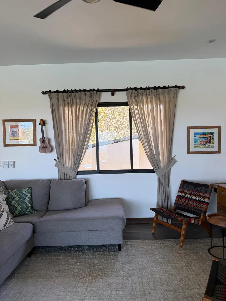 Living room with grey sectional sofa, sand pleated drapes with tie-backs, and tropical tree view.