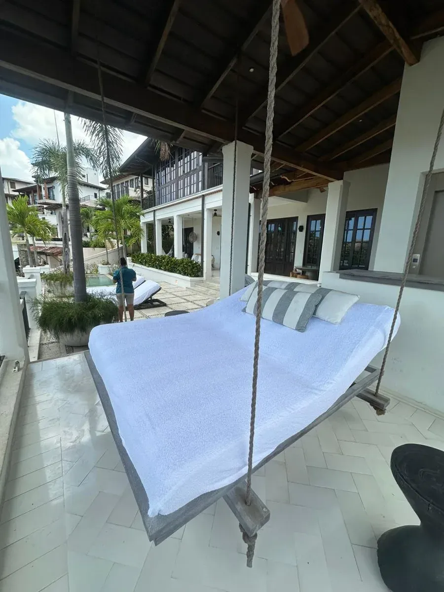 Covered terrace with hanging daybed on braided ropes, striped pillows, and colonial-house view.