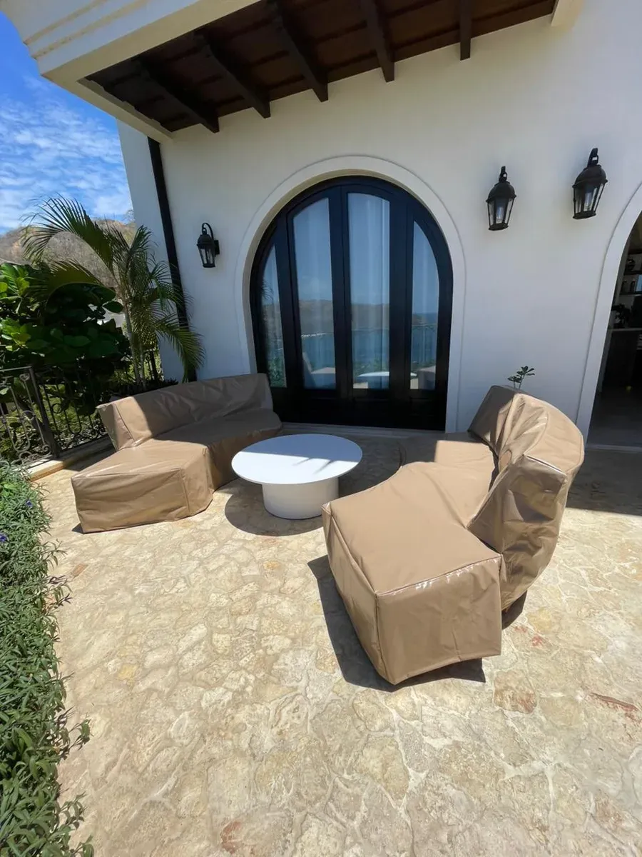 Patio with covered sofa and armchairs facing an arched French door on a colonial white facade.