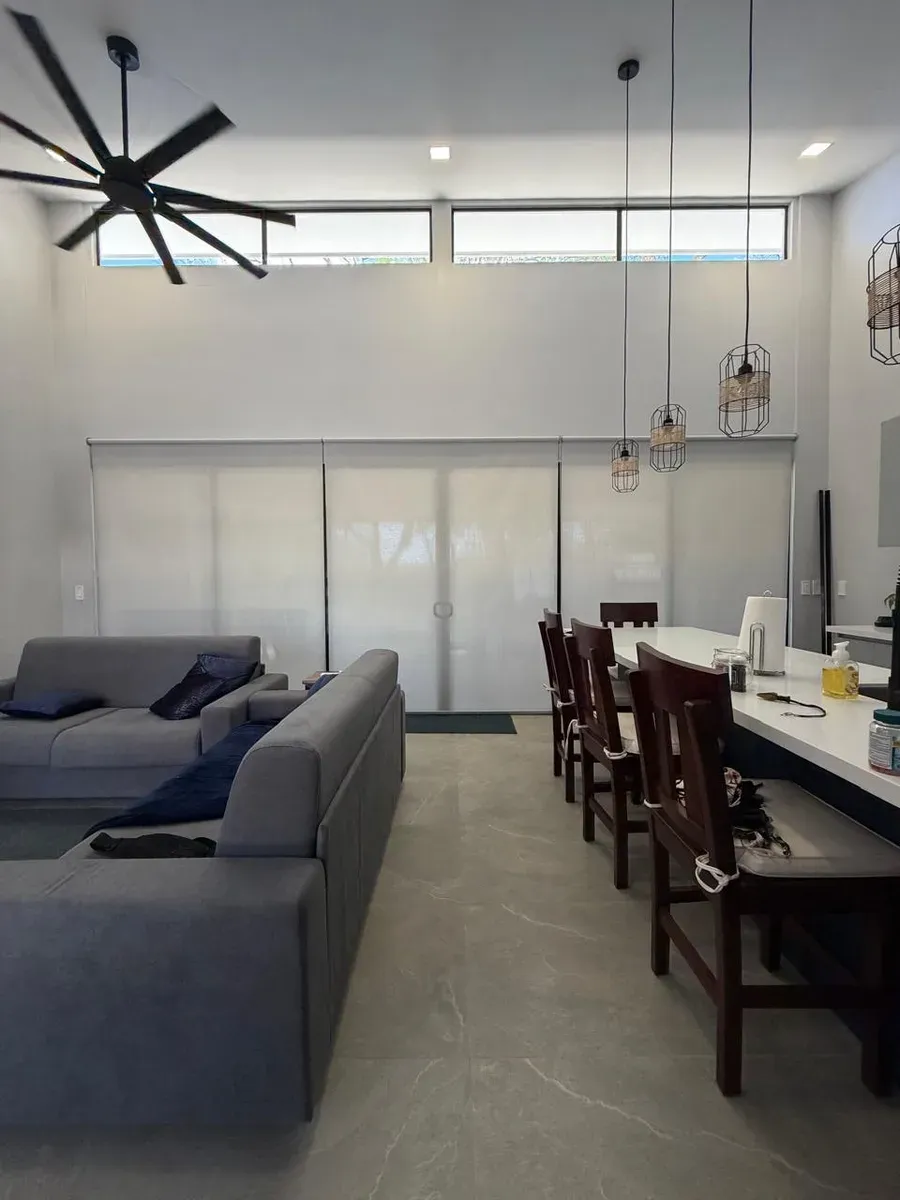 Open-plan room with double-height screen shades, industrial fan, and white bar with dark stools.