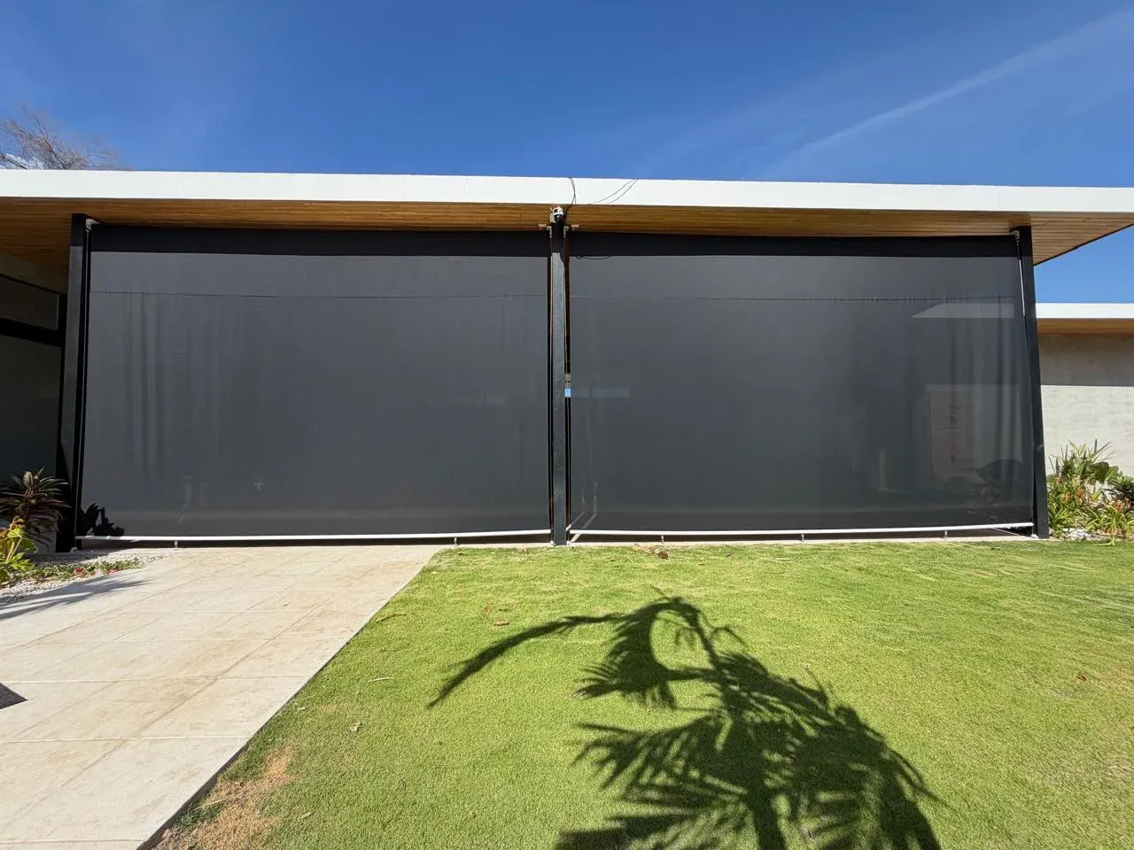 Facade with two charcoal exterior screen roller shades over a lawn with a palm-tree shadow.