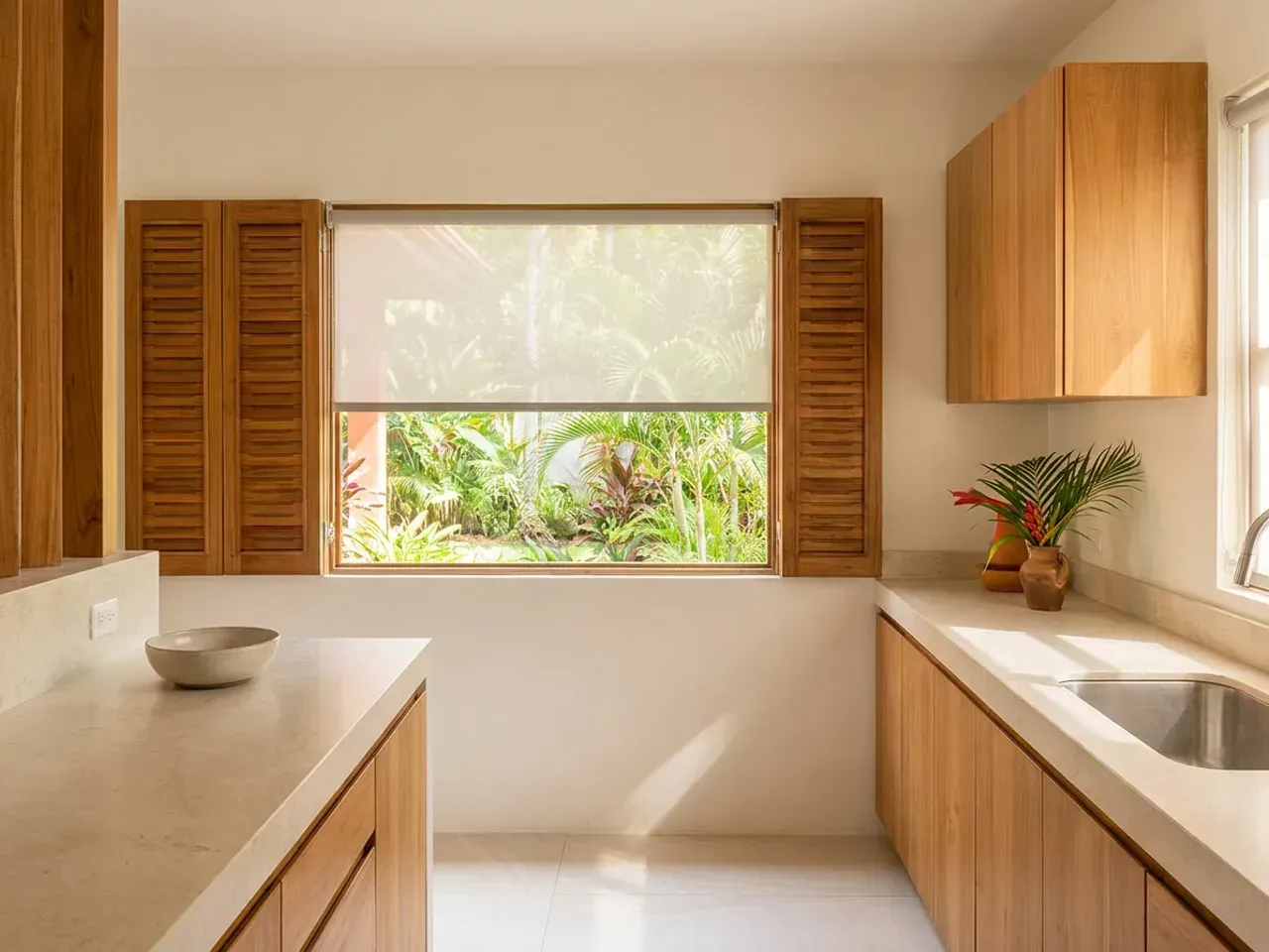 Cocina de teca clara con encimera de piedra lisa y Sun Screen bajado sobre la ventana, con vegetación tropical insinuándose a través del tejido.